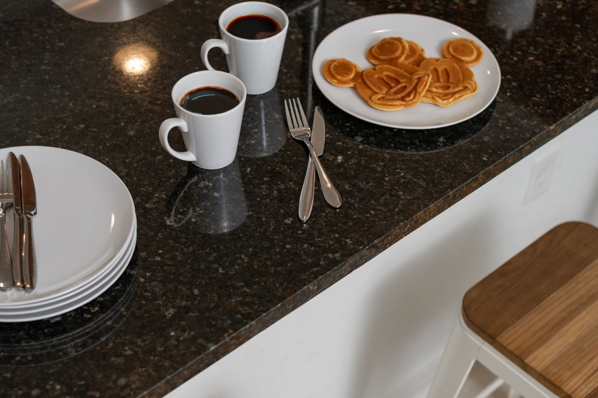 A kitchen counter with a plate of pancakes, two cups of coffee, and a fork and knife set.