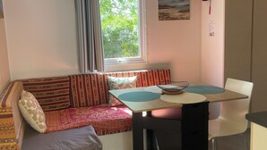 Une pièce de taille modeste, dotée d’une fenêtre, d’un canapé agrémenté de coussins à motifs, d’une table à manger avec un set de table et d’une chaise blanche.