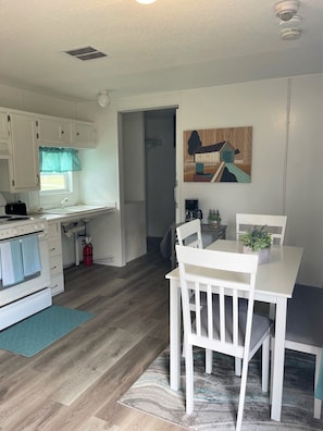 A compact kitchen with white cabinets, a stove, and a sink. There's a dining area with a table and chairs, a painting on the wall, and a fire extinguisher.