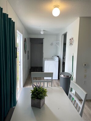 A room with a dining table, chairs, a potted plant, a washing machine, and a trash bin.