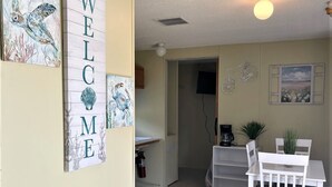 A room with a white table and chairs, a trash bin, a welcome sign, and a door leading to another room. - Sugar Valley RV Resort (Van Vleck)