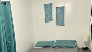 A bedroom with a bed, teal curtains, two wall-mounted leaf-patterned decorations, a bedside table with a lamp, and a nightstand.