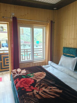 A room with a bed, a window with curtains, and a view of a building and greenery outside.