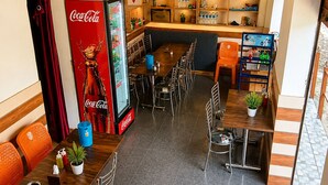 A restaurant with a Coca-Cola refrigerator, wooden tables and chairs, and shelves with various items. - Yamuna pag pinewood hotel  (Rajgarhi)