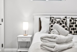 Una cama bien hecha con una pila de toallas blancas, una mesita de noche con una lámpara y almohadas estampadas.