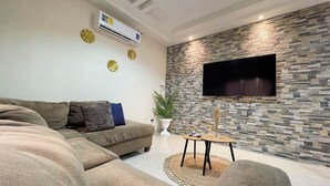 Un soggiorno con un divano componibile, una TV a schermo piatto montata su una parete con motivo in pietra e un condizionatore d'aria a soffitto.