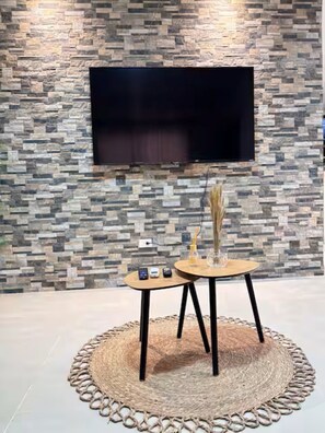 Una sala de estar moderna con una pared con diseño de piedra, un televisor de pantalla plana montado sobre una mesa redonda de madera con dos sillas, y una alfombra tejida en el suelo.