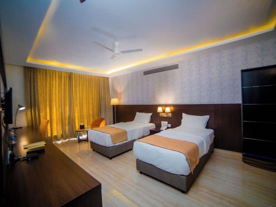 A hotel room with two beds, a desk, a chair, a TV, and a ceiling fan.