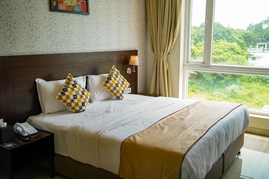 Une chambre d’hôtel avec un grand lit, deux coussins décoratifs, une lampe de chevet, une table de chevet avec un téléphone et une fenêtre donnant sur un paysage verdoyant.