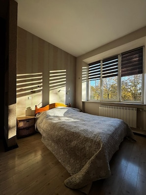 Un dormitorio con cama, mesita de noche, ventana con persianas y vistas a árboles.