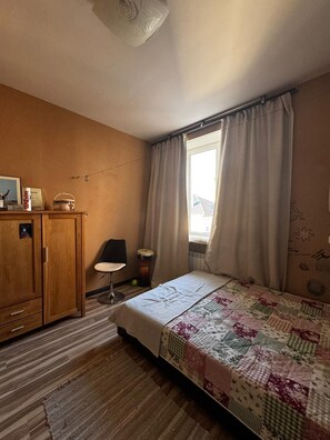 A bedroom with a wooden wardrobe, a bed with a quilt, a chair, a lamp, and a window with curtains. - Narva Promenade (Narva)