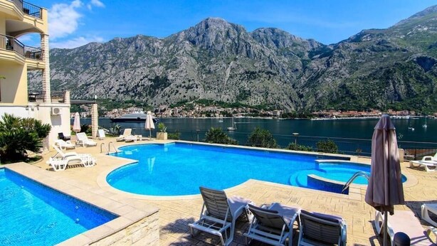 A swimming pool with lounge chairs and umbrellas, surrounded by mountains and a body of water. - The Breathtaking Views - Kotorscapes II (Muo)