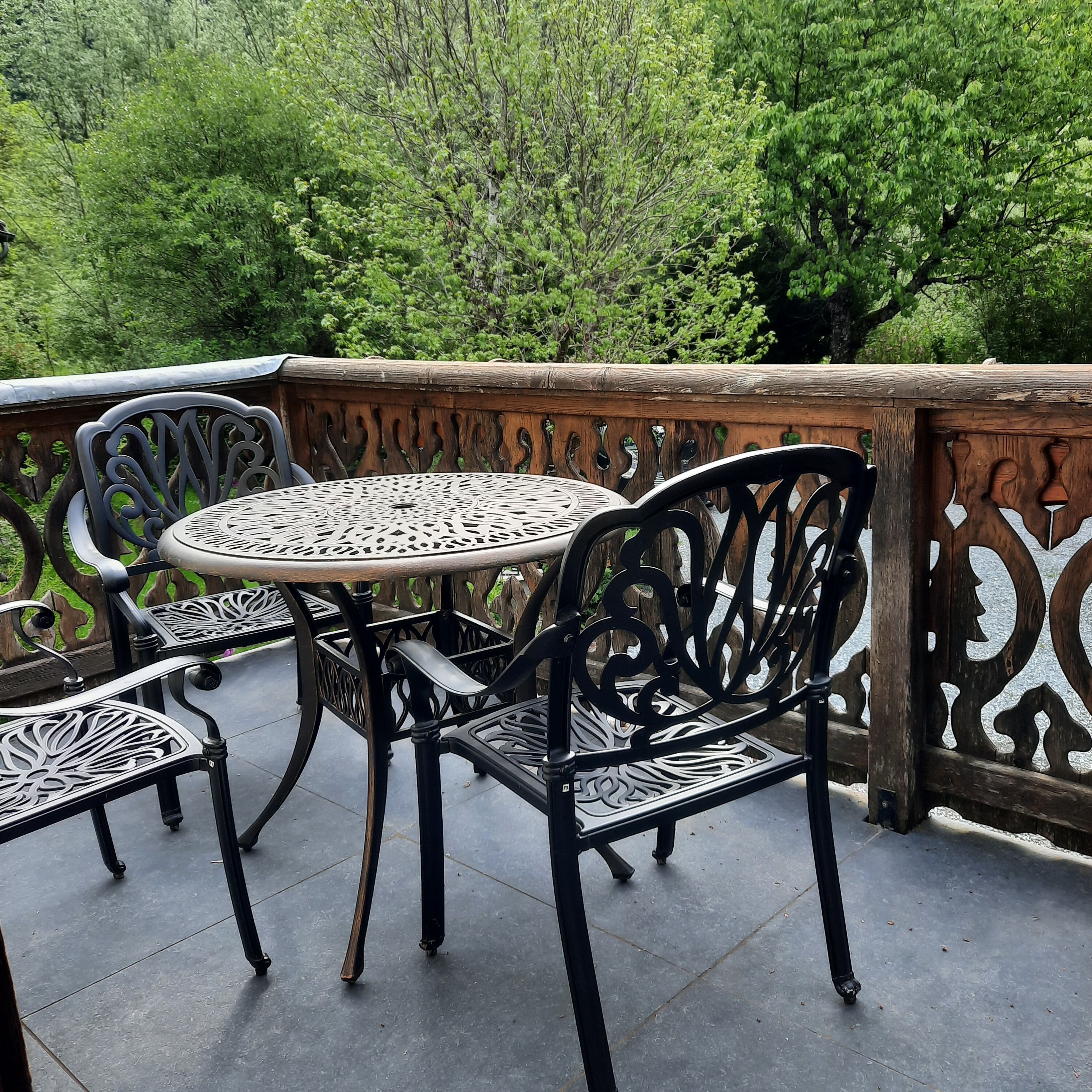 A round table with chairs, a wooden railing, and a view of green trees.