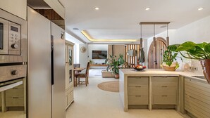 Una cocina moderna con electrodomésticos integrados, un comedor con mesa redonda y una sala de estar con televisor y plantas.