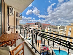 Un balcón con mesa y sillas de madera, una piscina y vistas de edificios cercanos.