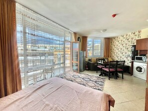 Una habitación con cama, zona de comedor, lavadora y vistas a edificios a través de amplios ventanales.