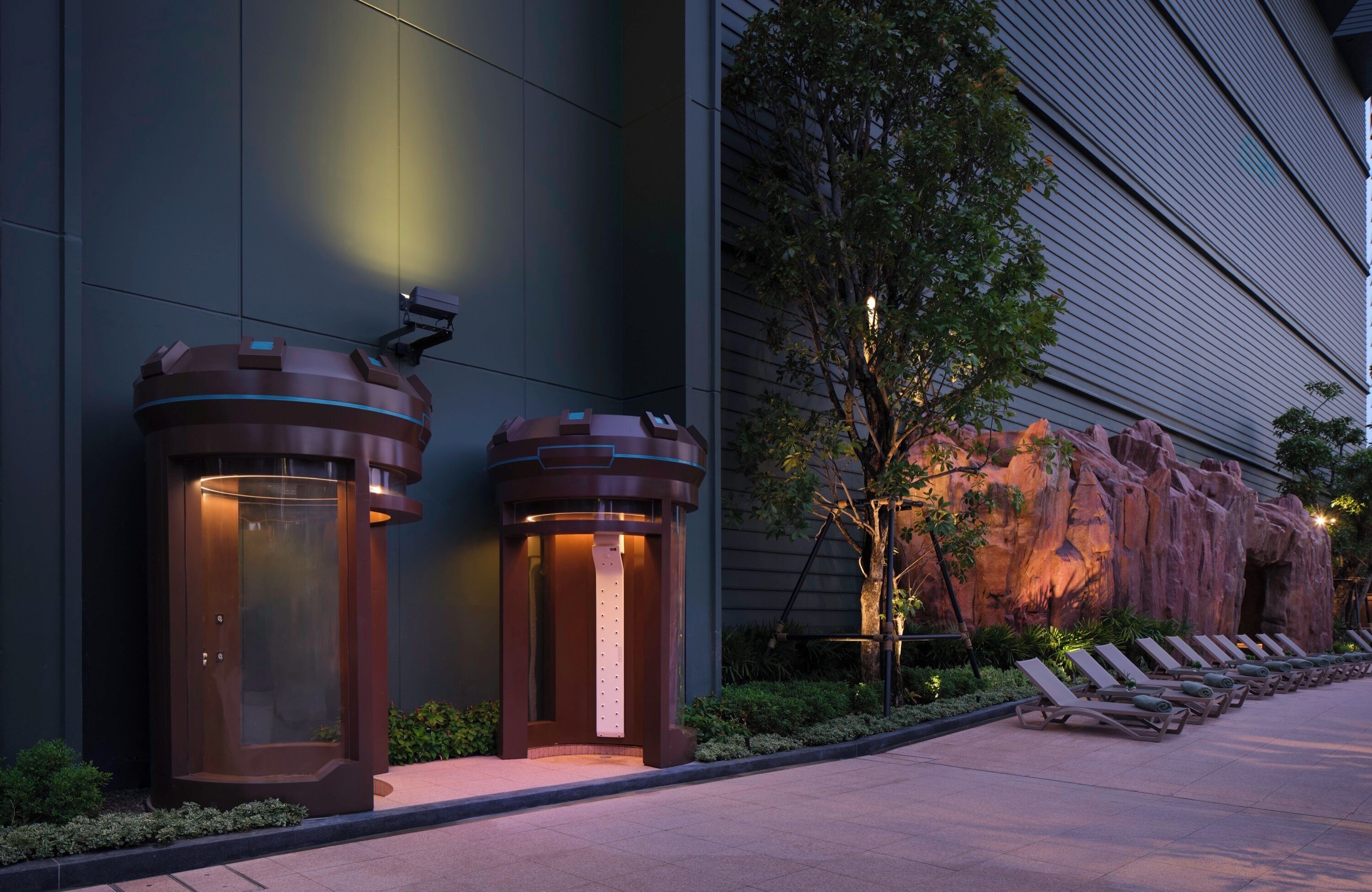 A modern building facade with two cylindrical structures, possibly security or information kiosks, and a landscaped area with lounge chairs and a rock formation.