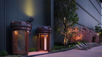 A modern building facade with two cylindrical structures, possibly security or information kiosks, and a landscaped area with lounge chairs and a rock formation.