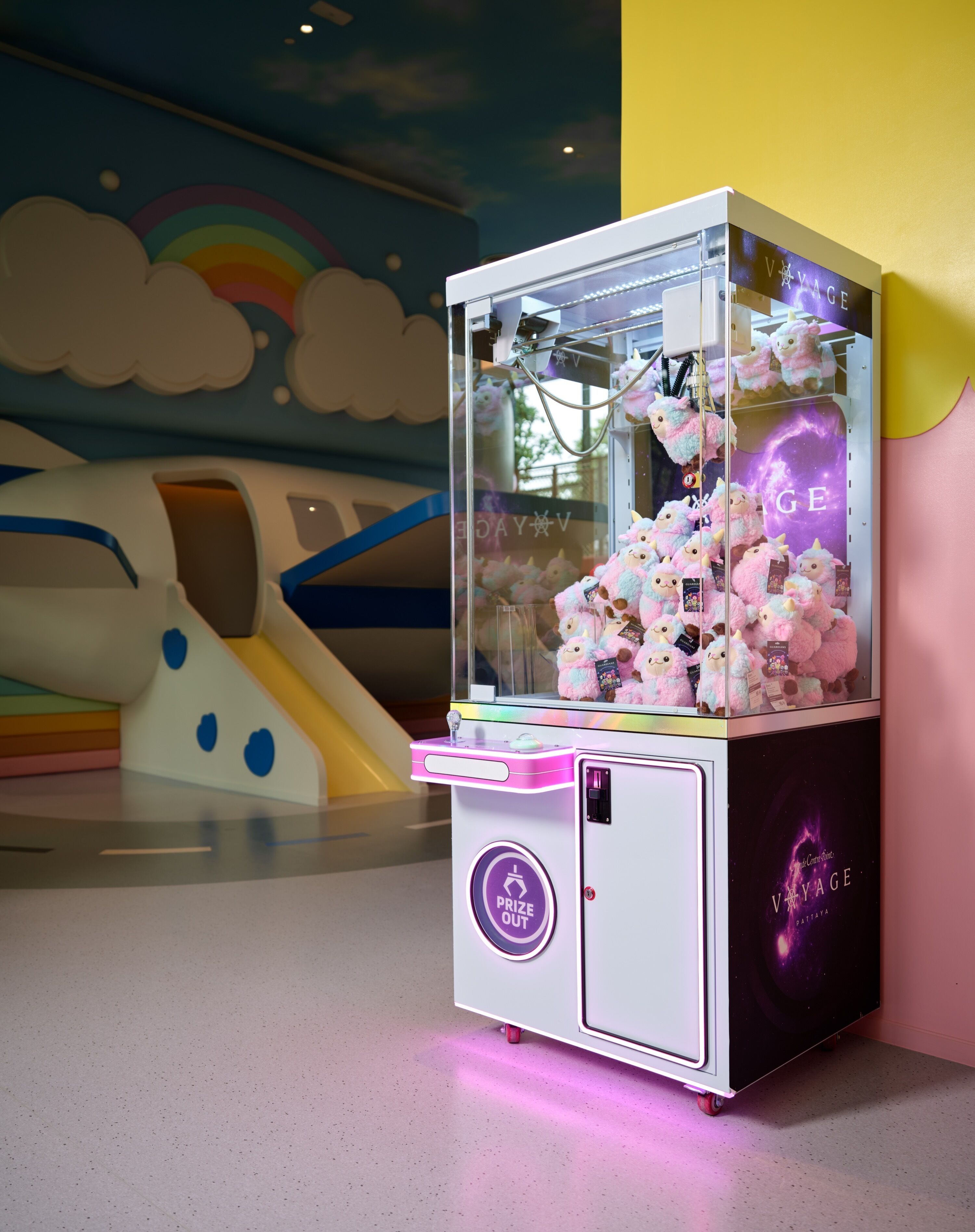 A claw machine with plush toys inside, located in a brightly lit indoor area with a colorful mural in the background.