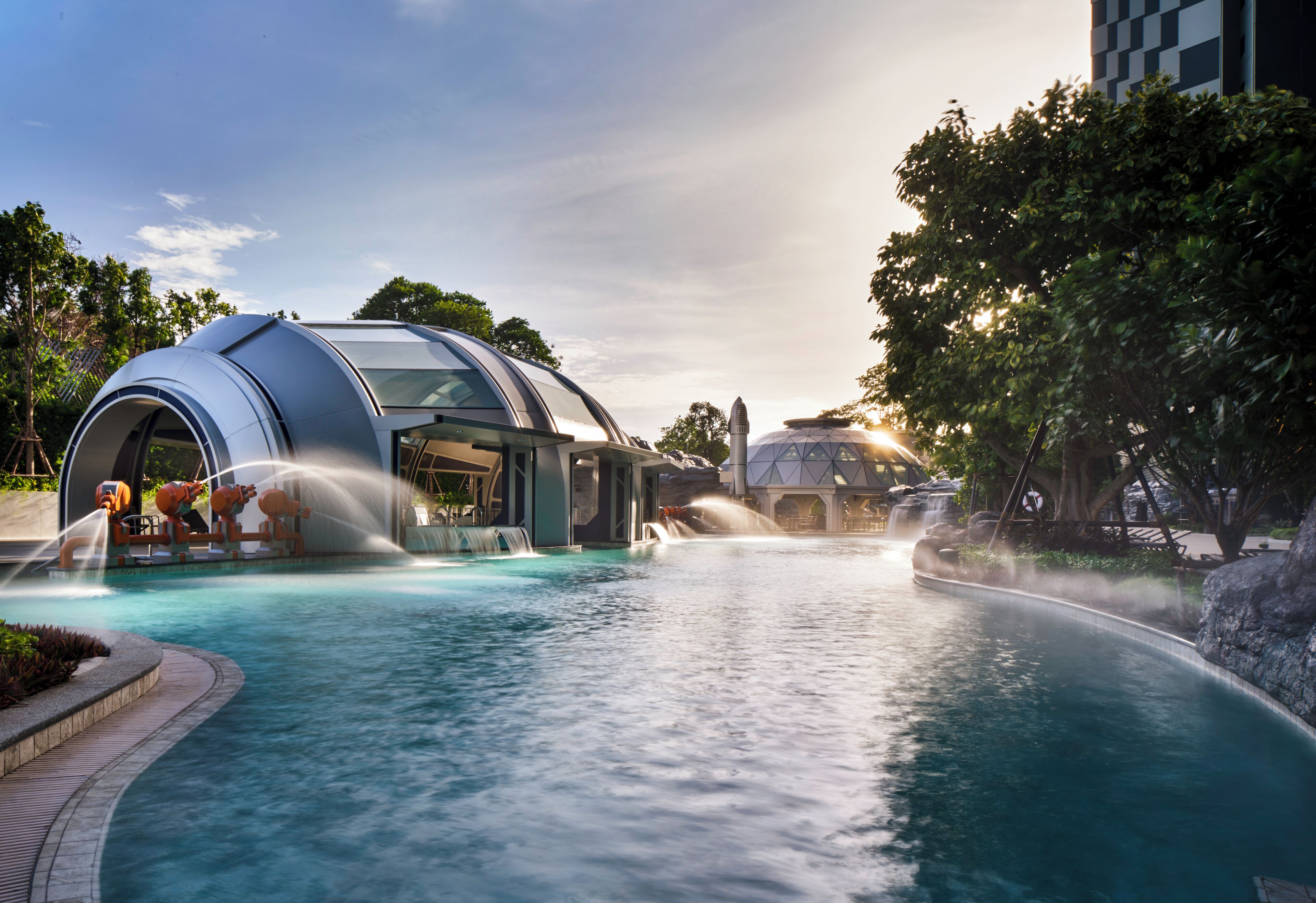 A modern pool area with a water feature, a covered seating area, and a building with a dome-like structure.