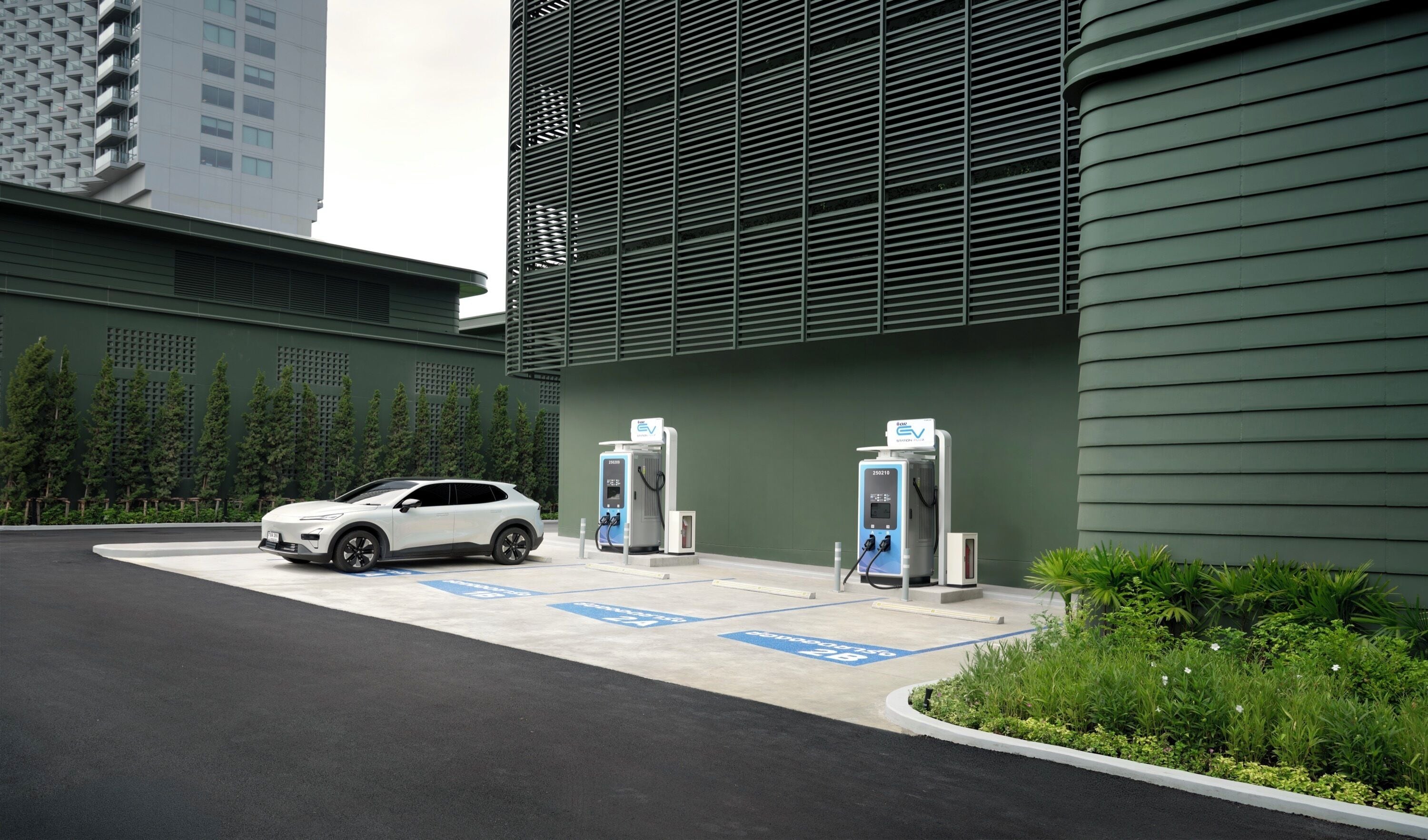 A parking area with electric vehicle charging stations, a white SUV, and a building with a green facade.