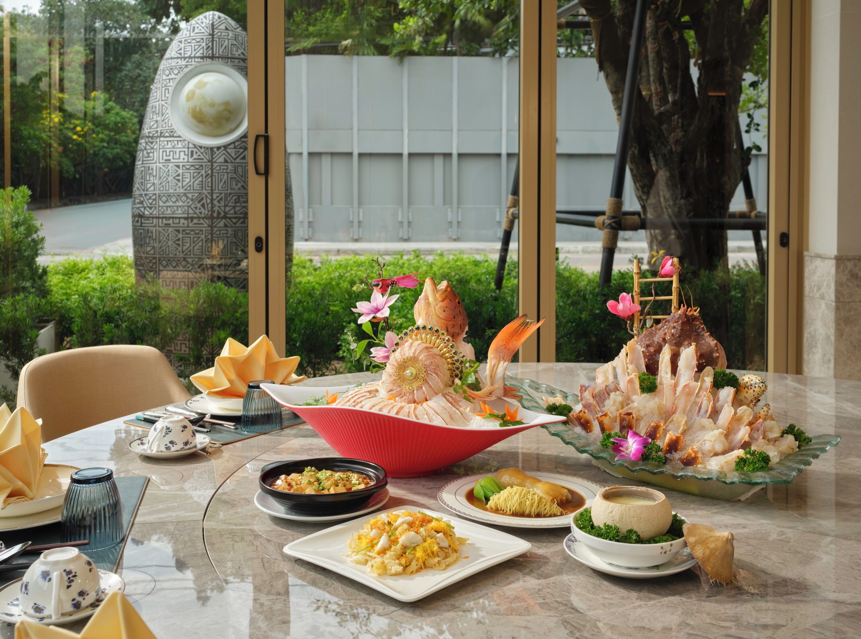 Una mesa de comedor con diversos platos, incluyendo mariscos y un tazón de sopa, ubicada en un espacio con vista a follaje y un árbol exterior.