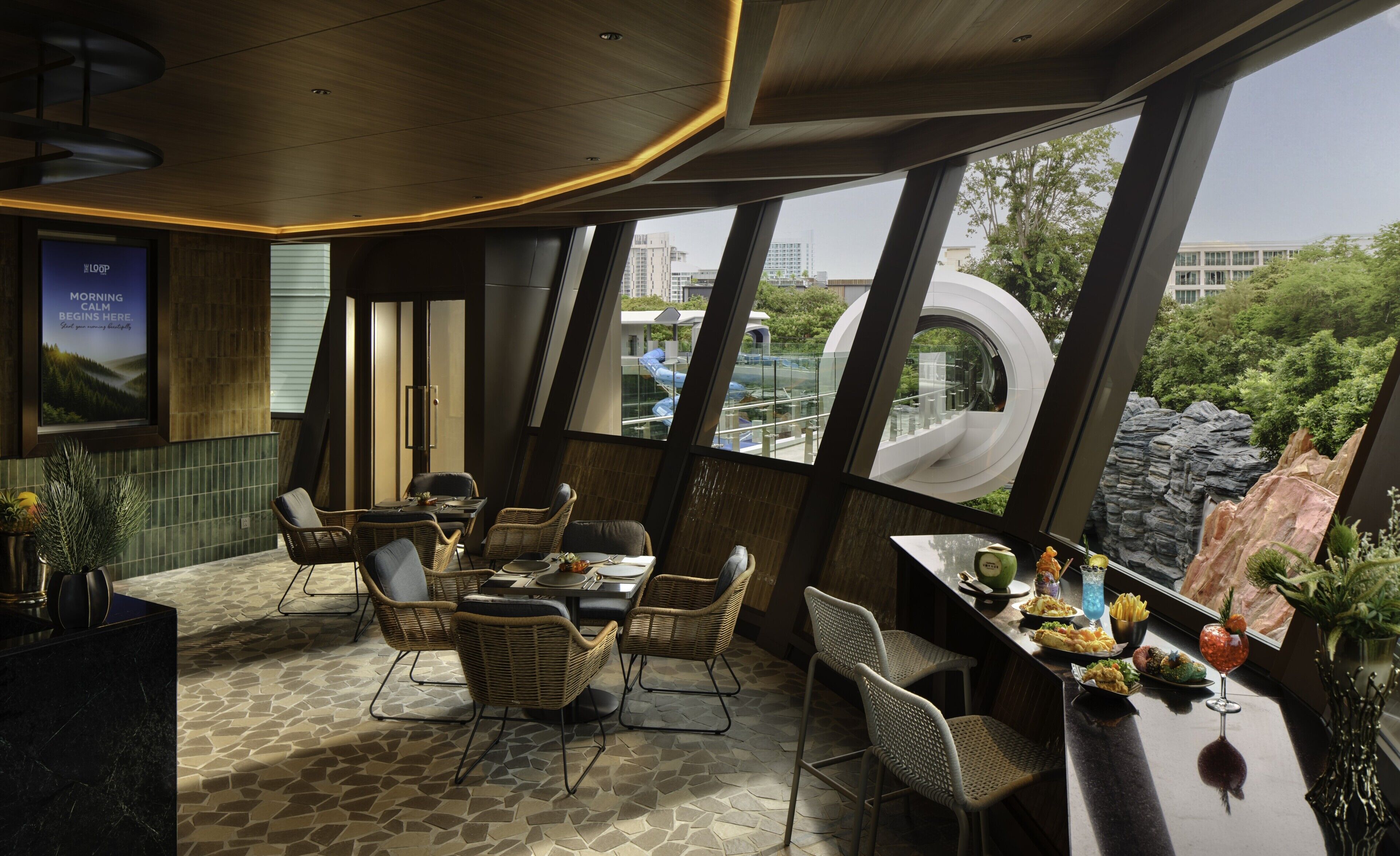 A restaurant with a modern interior, featuring a stone-tiled floor, wicker furniture, and large windows offering a view of the cityscape and greenery.