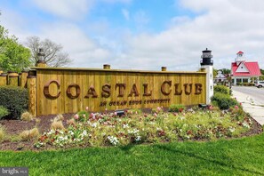 Ein Holzschild für Coastal Club in der Ocean Ridge Community, im Hintergrund ein Leuchtturm und ein Gebäude mit rotem Dach.