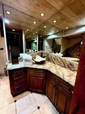 Une salle de bain avec un comptoir en marbre, des meubles en bois, un grand miroir et des spots encastrés au plafond.