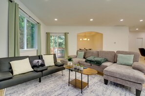 Ein modernes Wohnzimmer mit Sofa, Sessel und Couchtisch, großen Fenstern mit Vorhängen und einer Pendelleuchte.
