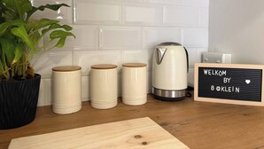 Une cuisine avec des carreaux blancs, une machine à café, trois pots à sucre, une plante en pot et un panneau d’accueil.