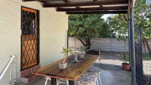 Une terrasse couverte avec une table en bois, une plante en pot et une porte ornée d’une grille métallique.