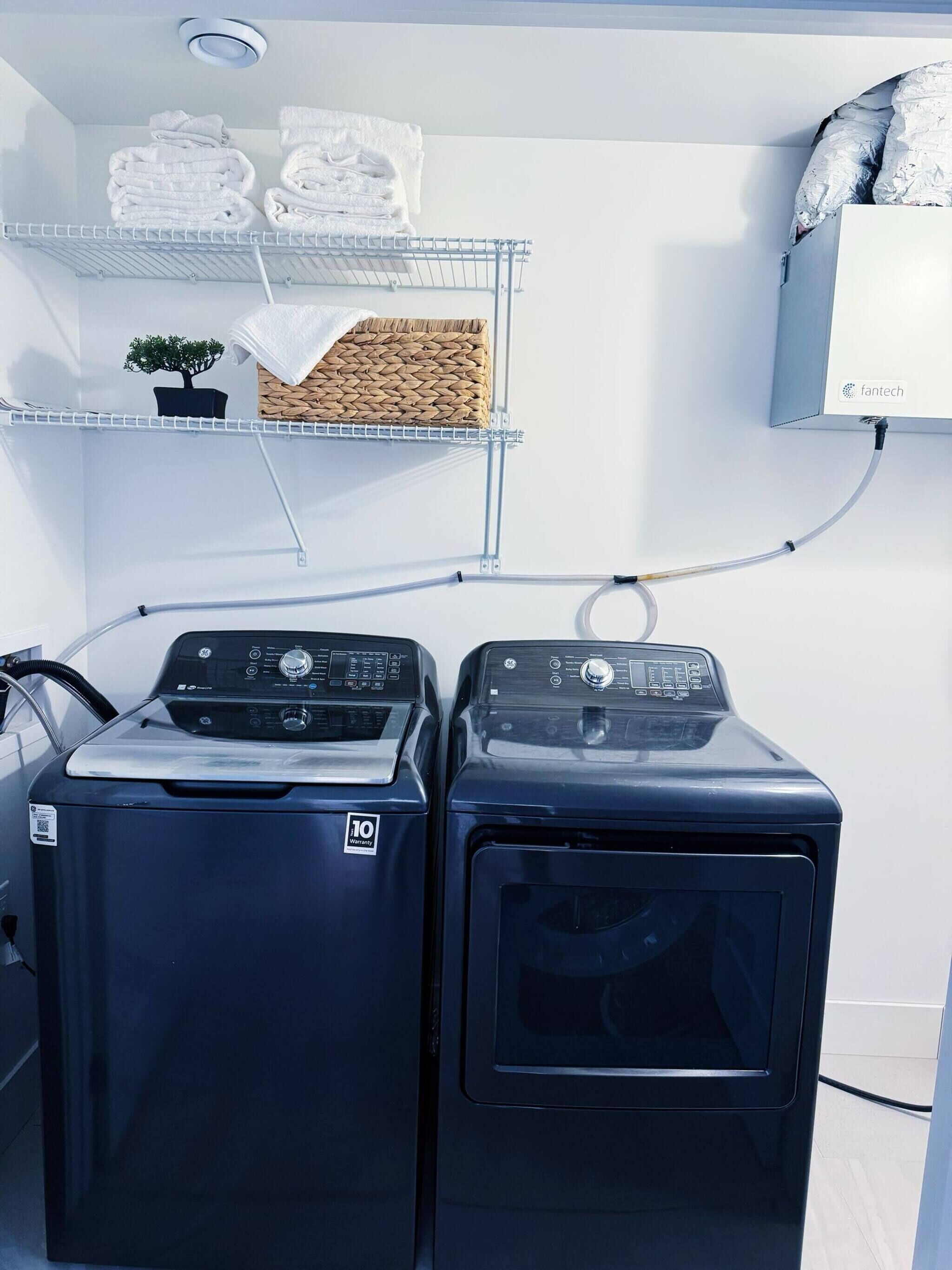 Un espace buanderie comprenant deux machines à laver à chargement frontal, une étagère avec des serviettes pliées et une petite plante en pot.