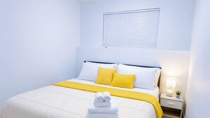 Ein ordentlich eingerichtetes Hotelzimmer mit Bett, Nachttisch, Lampe und einem Fenster mit Jalousien.