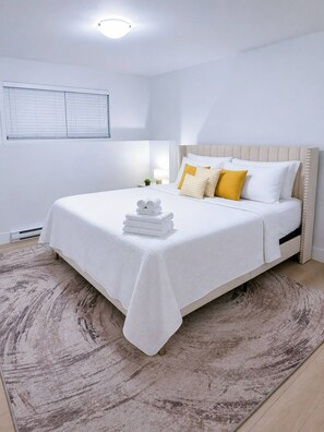 A neatly made bed with a white bedspread and pillows, a small bedside table with a lamp, and a large window with blinds. - Stylish 3-bedroom Full House Near Magnetic Hill (Moncton)
