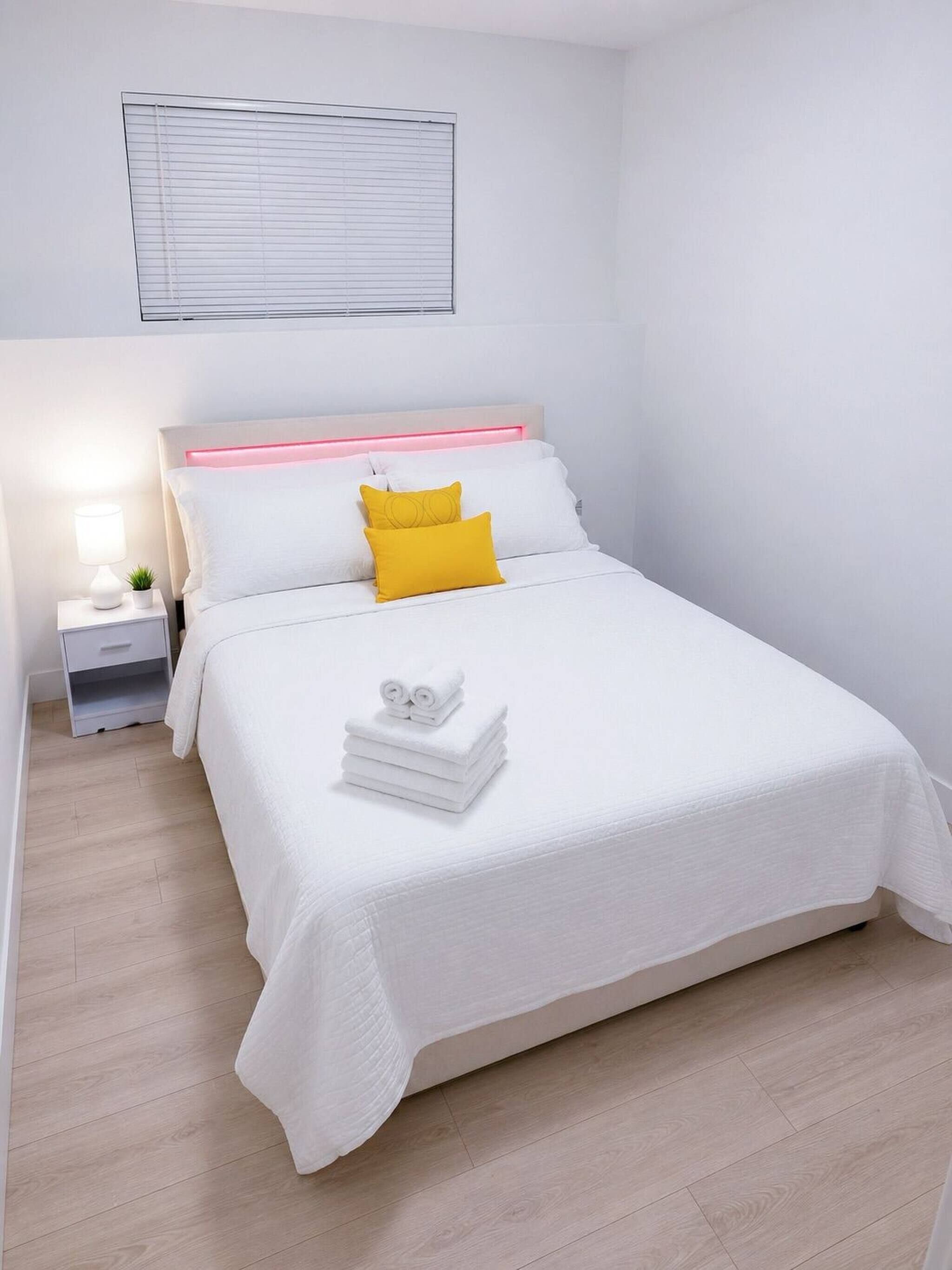 Ein ordentlich eingerichtetes Hotelzimmer mit einem Bett, einem Nachttisch, einer Lampe und einem Fenster mit Jalousien.