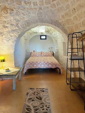 A room with a bed, a desk with a lamp, a small table with a bowl of fruit, and a hanging shelf. - Authentic trulli centrally in Valle d'Itria (Ceglie Messapica)