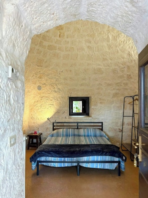 A bedroom with a stone wall, a bed with striped bedding, a small table with a red cup, a window with a view, and a metal ladder. - Authentic trulli centrally in Valle d'Itria (Ceglie Messapica)