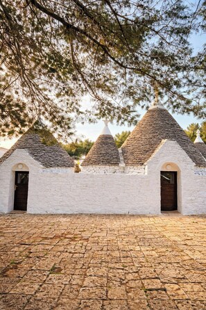 A building with three conical roofs and white walls, a paved ground, and trees in the background. - Authentic trulli centrally in Valle d'Itria (Ceglie Messapica)