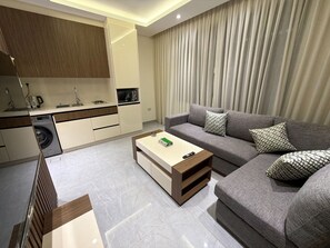 A modern living room with a grey sofa, a coffee table, and a kitchen area with a washing machine. - 104 JRE apartment with stunning boulevar (Amman)