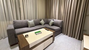 Ein modernes Wohnzimmer mit einem grauen Ecksofa, einem Holztisch und einem großen Fenster mit transparenten Vorhängen.