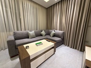 Ein modernes Wohnzimmer mit einem grauen Ecksofa, einem Holztisch und einem großen Fenster mit transparenten Vorhängen.