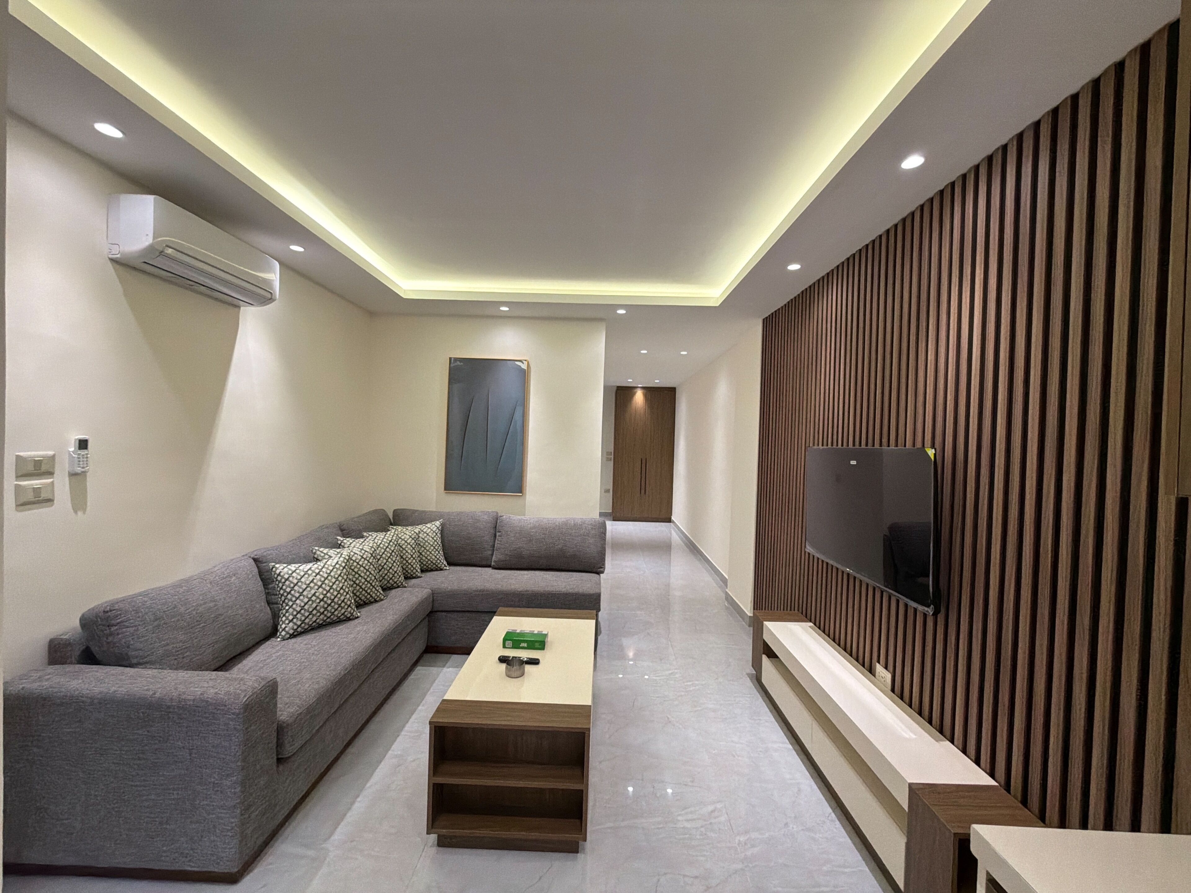 A modern living room with a sectional sofa, a coffee table, a flat-screen TV mounted on a wooden panel wall, recessed ceiling lighting, and a hallway leading to a door.