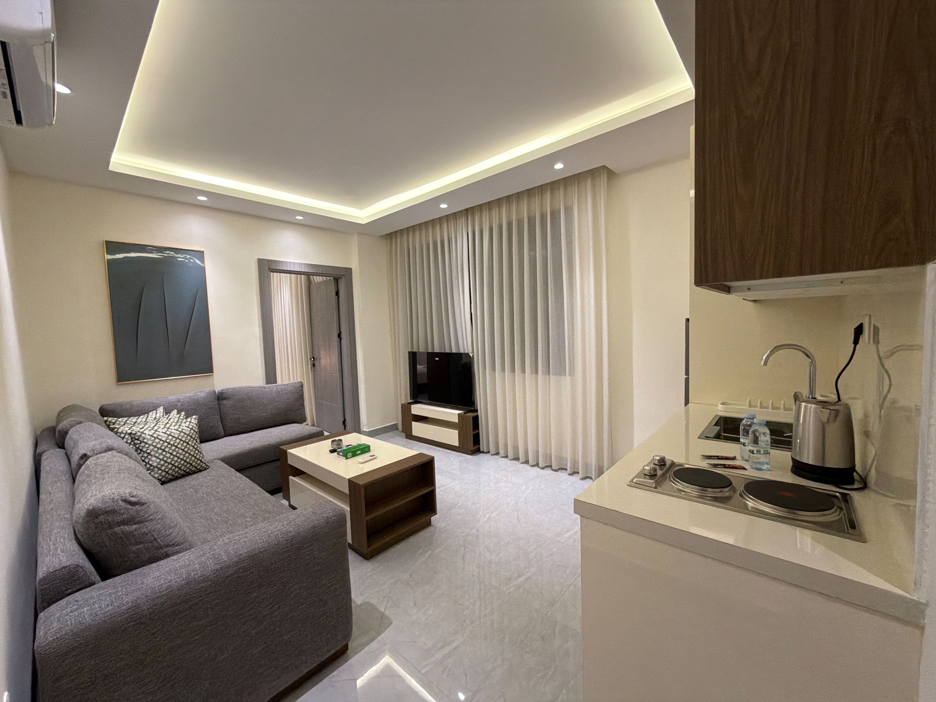 Una sala de estar moderna con sofá, mesa de centro, televisor y una zona de cocina con fregadero y hervidor.