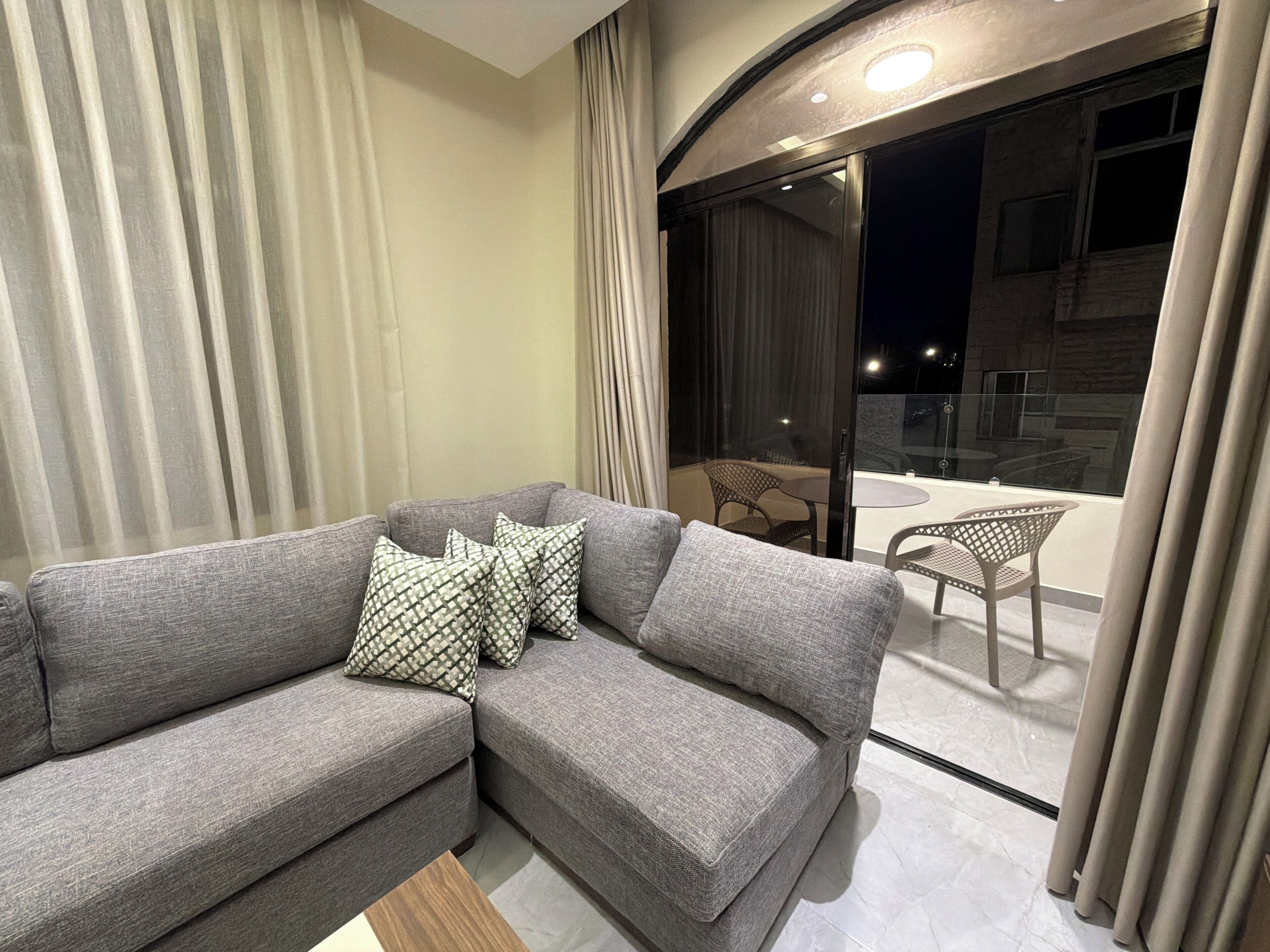 A modern living room with a grey sofa, patterned cushions, a wooden coffee table, and a glass door leading to a balcony with chairs.