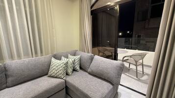 A modern living room with a grey sofa, patterned cushions, a wooden coffee table, and a glass door leading to a balcony with chairs.