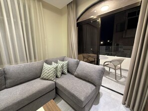 Ein modernes Wohnzimmer mit einem grauen Sofa, gemusterten Kissen, einem Holztisch und einer Glastür, die auf einen Balkon mit Sitzgelegenheiten führt.