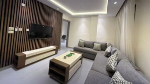 A modern living room with a sectional sofa, a flat-screen TV mounted on a wooden panel wall, a coffee table, and a built-in bench. - 301 JRE 2Bedr rooftop with beautiful view (Amman)