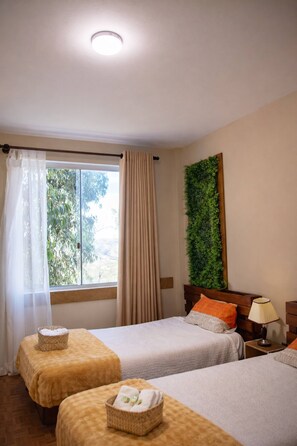 Et hotelværelse med to senge, et vindue med gardiner, en grøn plante på væggen og et natbord med en lampe.