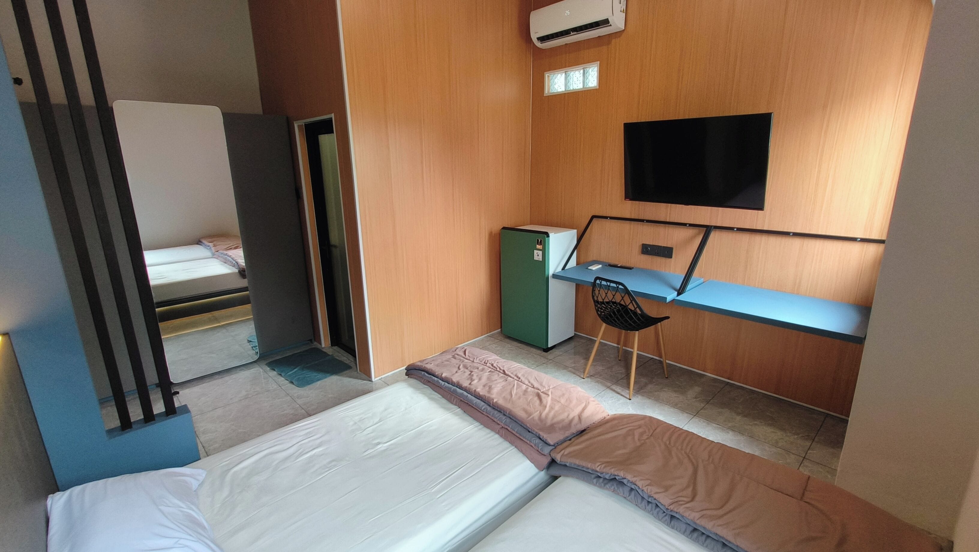Ein kleines Zimmer mit einem Bett, einem Schreibtisch mit Stuhl, einem kleinen Kühlschrank und einem Fernseher an der Wand.